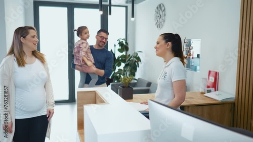 A pregnant woman with her husband and little daughter visits a medical clinic for a gynecological examination. Friendly female hospital receptionist talking to clients at the reception desk in a medic photo