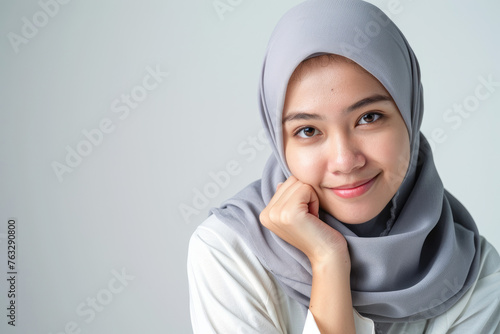 A beautiful young Malay woman wearing a hijab, smiling and posing for the camera with her hand on her chin against a white background 