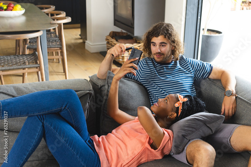 A diverse couple relaxes on gray sofa at home, using a smartphone