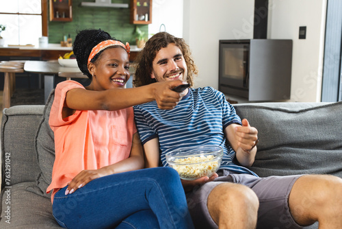 A diverse couple enjoys a cozy moment on the sofa at home, eating popcorn and watching television