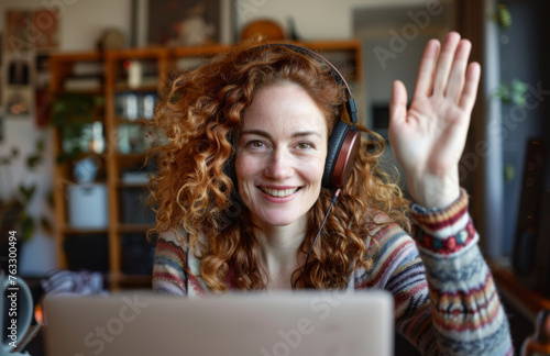 An attractive woman with red hair in headphones greeting with a smile at her laptop