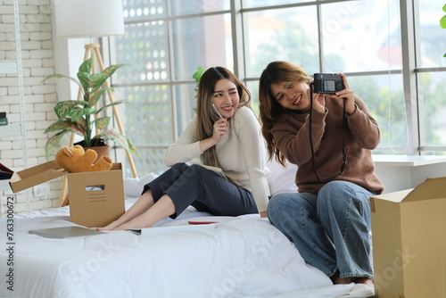 Two Asian female roommates sit on the bed together, friend holding a camera taking a selfie with a friend in the bedroom of an apartment. photo