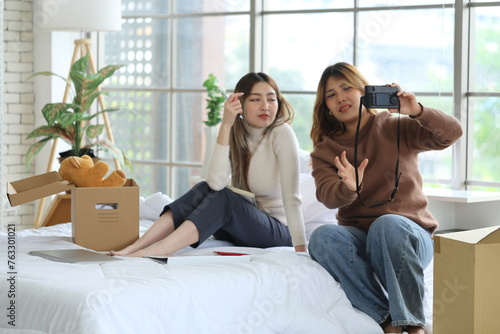 Two Asian female roommates sit on a bed together, one holds a camera and takes a selfie together, while fellow writers document a good relationship in an apartment room