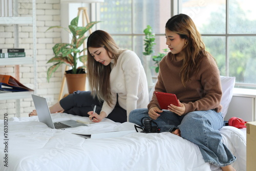 Two young Asian female friends, freelance bloggers, roommates, sit on a bed and write travel reviews together, use laptops and make reports for websites together, and live in an apartment. photo