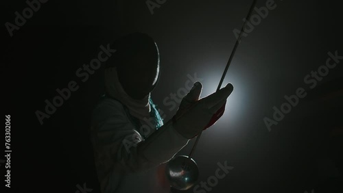 Medium footage of foilsman in epee mask holding rapier and looking at its blade while standing against lightMedium footage of foilsman in epee mask holding rapier and looking at its blade while standi photo