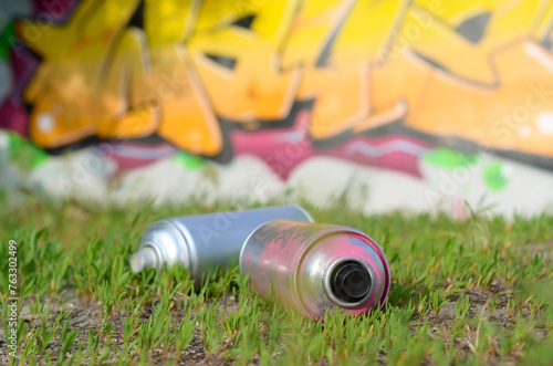 Several used spray cans with paint and caps for spraying paint under pressure on grass near the painted wall in colored graffiti drawings
