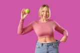 Pretty young woman in loose jeans with apple on pink background. Weight loss concept