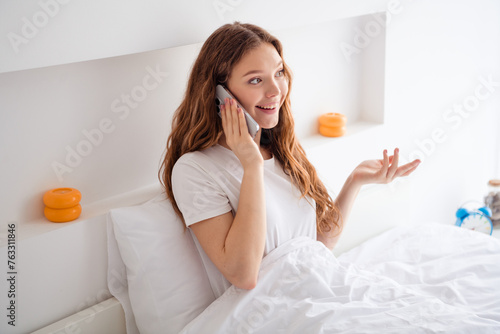 Photo of charming cheerful girl sit bed under blanket speak communicate telephone pastime morning bedroom indoors