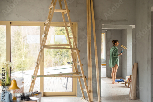 Young woman paints walls while making repairment of a new house. Standing with paint roller and looks out. Creative process of home renovation and repair concept