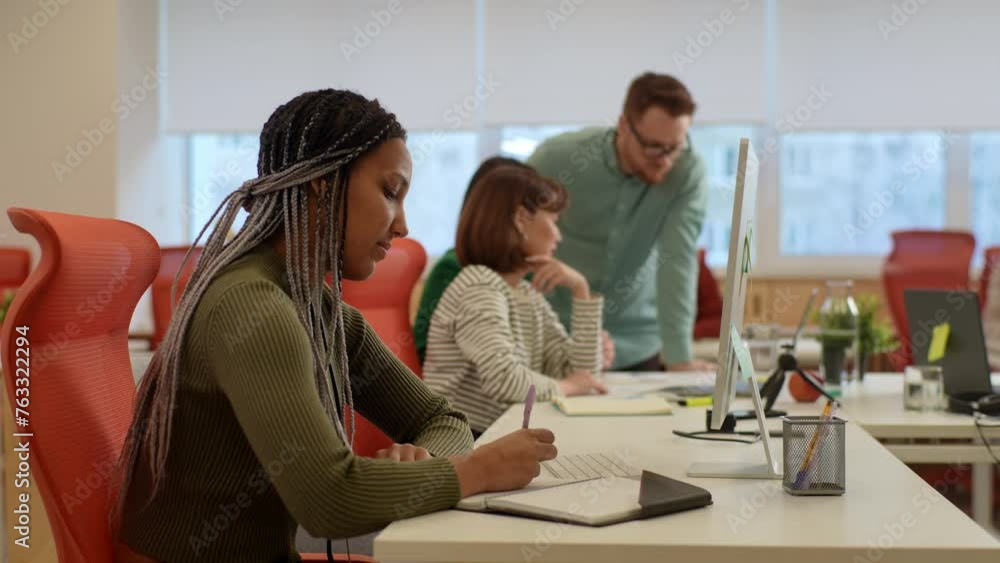 Focused African female manager prepare financial report making presentation writes notes or corrections, doing paperwork sitting at modern workplace in office. Businesspeople negotiating on background Focused African female manager prepare financial report making presentation writes notes or corrections, doing paperwork sitting at modern workplace in office. Businesspeople negotiating on background
