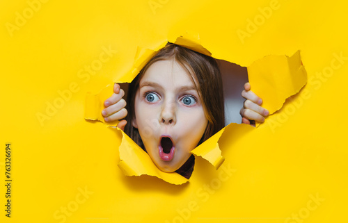 Funny little red-haired child girl peeping through hole on yellow paper. The concept of surprise, fear, fright, joyful mood from what he saw. Discounts, sales, surprise. Copy space.