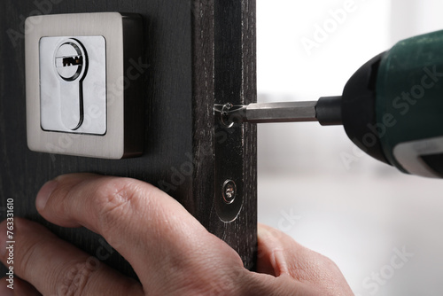 Handyman with electric screwdriver repairing door lock indoors, closeup photo