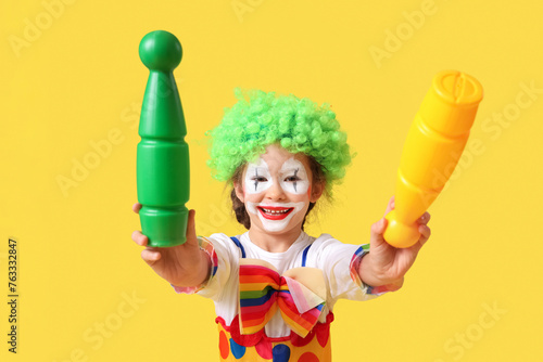 Funny little girl in clown costume with juggling clubs on yellow background. April Fools' Day celebration