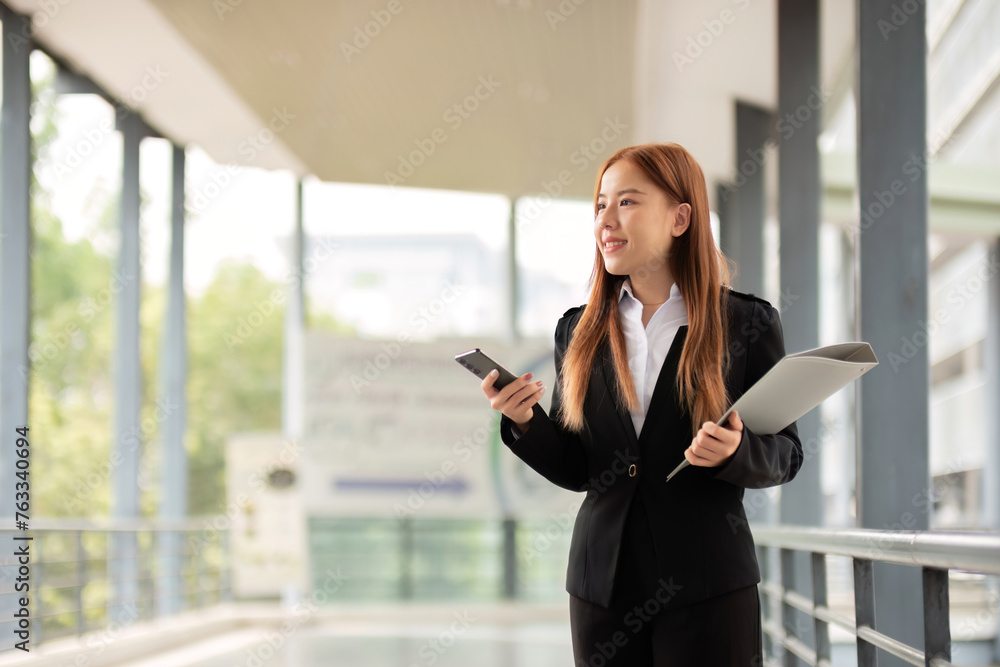 Fototapeta premium Portrait of young Asian businesswoman in suit talking on smartphone while standing in front of modern office building business concept and people