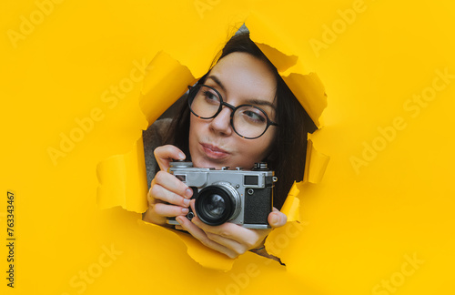 A young paparazzi girl in glasses with a rare SLR camera looks out from her hiding place and carefully looks at what is happening. Yellow paper hole. Tabloid press.Looking for a story for stock photos