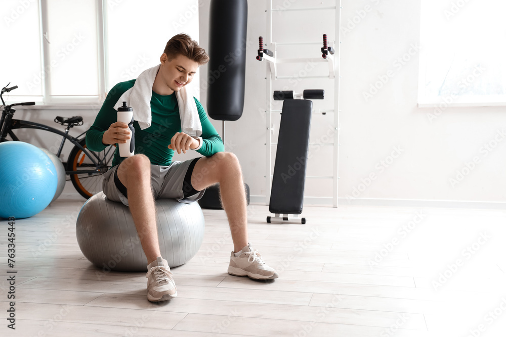 Fototapeta premium Sporty young man with water bottle looking at smartwatch on fitball in gym
