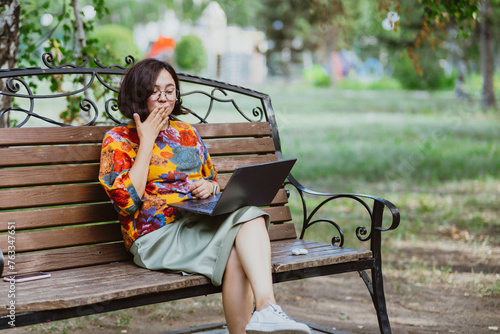 Asian woman blowing kiss in video call while sitting with laptop on bench in green park. Adult woman outdoors communicating with loved ones via video call on laptop