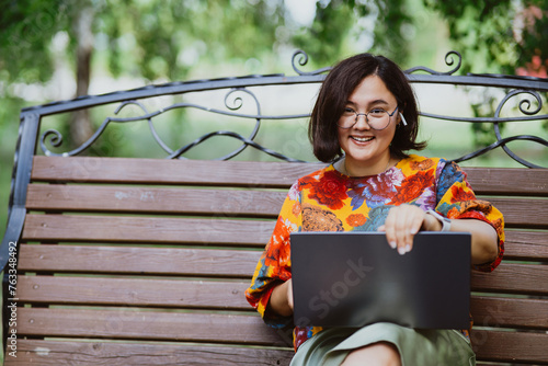 Smiling Asian woman working on her laptop while sitting on a park bench Freelancer lifestyle depicted by a woman working on technology in a quiet park