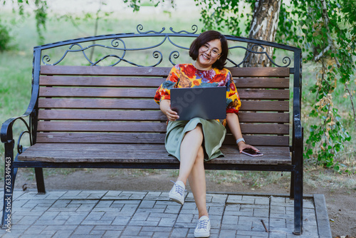 A cheerful woman works on her laptop while sitting on a park bench surrounded by birch trees. Nature meets technology with a fashionable woman using her laptop outdoors on a sunny day