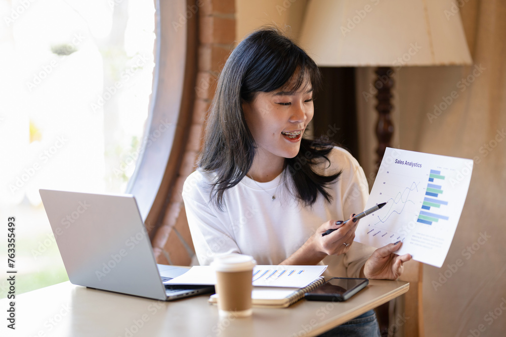 Young Asian businesswoman presents sales analysis charts during an online meeting in a coffee shop area. Young Asian businesswoman presents sales analysis charts during an online meeting in a coffee shop area.
