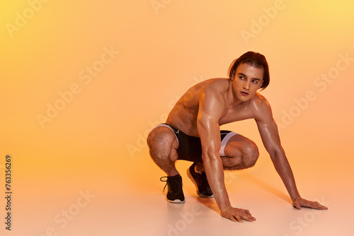 attractive shirtless man in black shorts exercising actively and looking away on orange backdrop