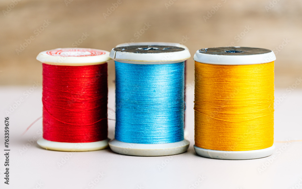 Three spools of thread in vibrant red, blue, and yellow stand in a row against a neutral background, illustrating the basic tools of sewing and textile arts. Three spools of thread in vibrant red, blue, and yellow stand in a row against a neutral background, illustrating the basic tools of sewing and textile arts.