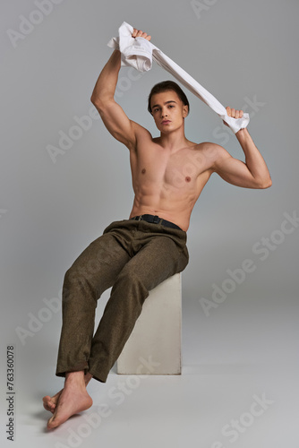 tempting shirtless man in brown pants sitting and looking alluringly at camera on gray backdrop photo