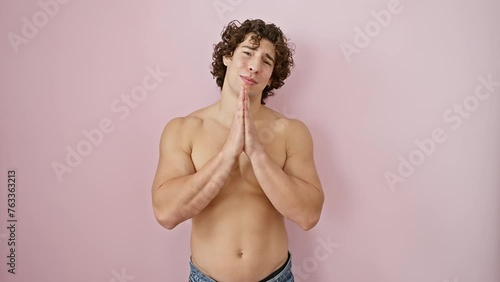 Handsome shirtless young hispanic man fervently praying, hands clasped, begging for forgiveness, amid hopeful thoughts, over a pink isolated background. photo