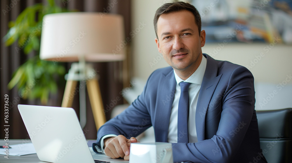 Portrait of a successful male businessman, banker, financial director sitting in the office at the table and using a laptop. Looking and smiling at the camera Portrait of a successful male businessman, banker, financial director sitting in the office at the table and using a laptop. Looking and smiling at the camera