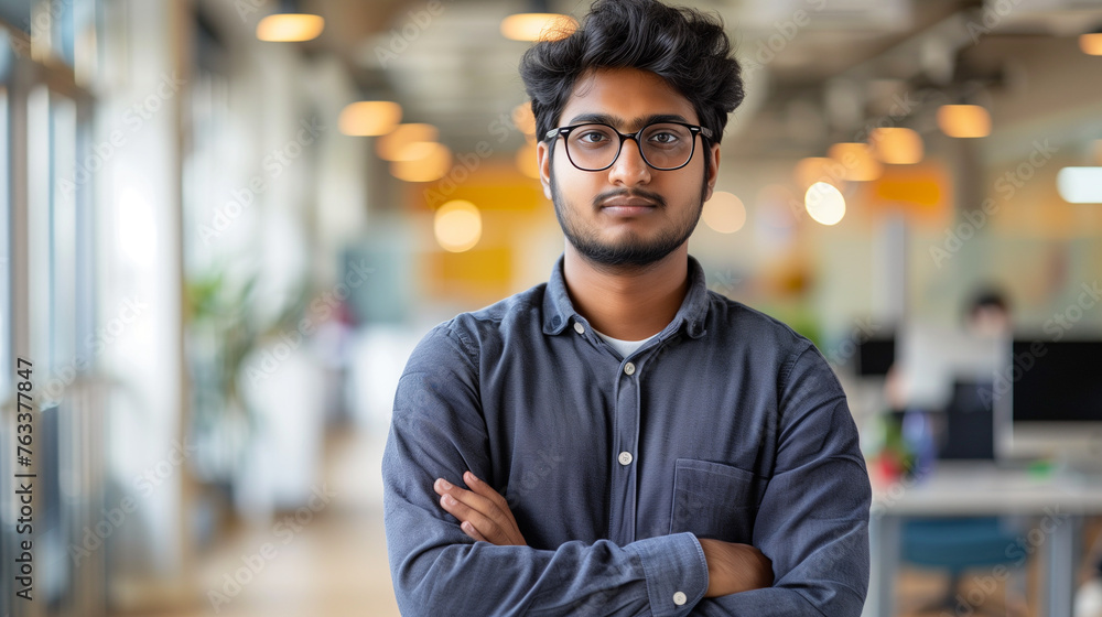 Portrait of a young successful Indian male programmer, developer standing in an office center, crossing his arms over his chest and looking seriously at the camera - obrazy, fototapety, plakaty 