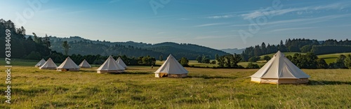 An outdoor meadow with white camping tents against a background of blue sky and green trees. © SHI