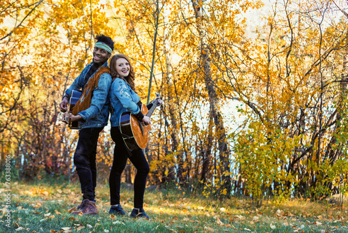 Mixed race married couple standing back to back, smiling and posing for the camera playing guitars during a fall family outing in a city park, spending quality time together; Edmonton, Alberta, Canada photo