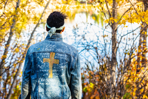 Close-up view from behind of a man wearing a jean jacket with hand-written Christian references during a fall outing in a city park, standing by a lake; Edmonton, Alberta, Canada