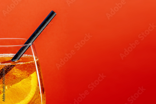 Detail of aperol spritz aperitif with orange slice, ice cubes and black drinking straw on bright red background with space for text.