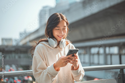 Beautiful young happy asian woman with headphones looking at mobile phone while standing in the city