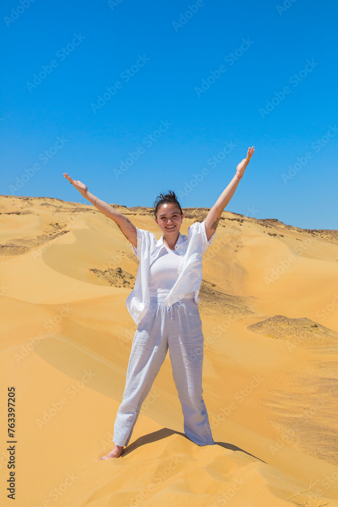 Young woman in white clothing stands with arms raised on the sand dunes of the White Desert Protected Area in Egypt; Egypt Young woman in white clothing stands with arms raised on the sand dunes of the White Desert Protected Area in Egypt; Egypt
