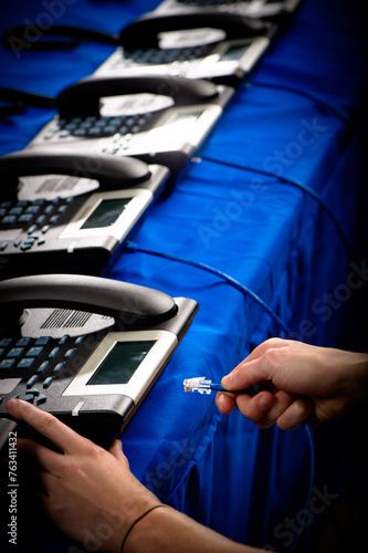 A hand plugs a phone cable into a telephone on a table full of phones in a call center photo