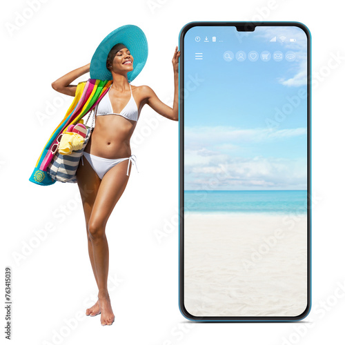 Happy girl in summer beach holiday wearing bikini and sun hat hold beach bag and towel, showing a big screen mobile phone isolated in white background, online shopping or booking sea vacation travels