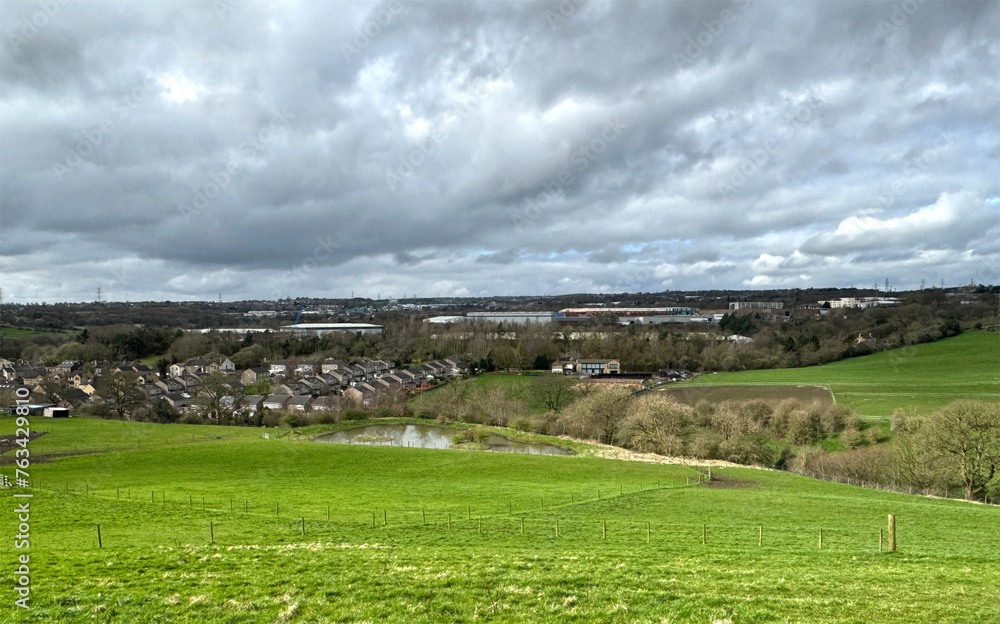 Obraz premium Landscape view from, Cliff Hollins Lane, with a large field, a lake, and distant houses, on a cloudy day near, Dewsbury, UK