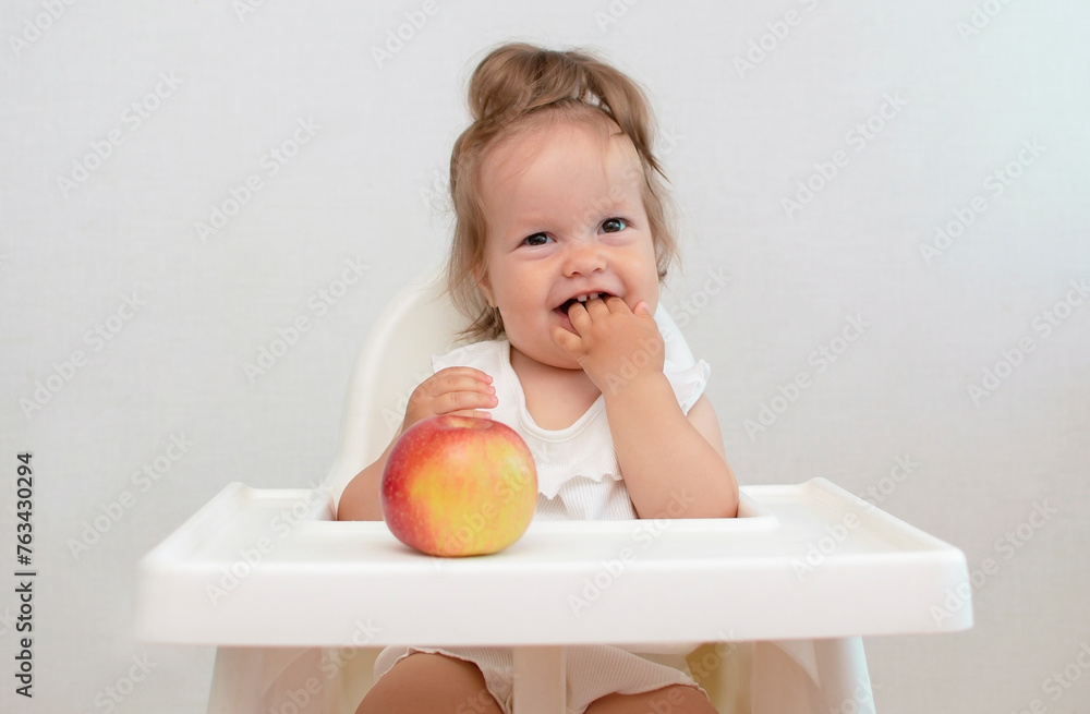 Small child sitting in high chair and eating big red apple. The girl eats healthy food Small child sitting in high chair and eating big red apple. The girl eats healthy food