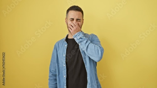 Hispanic guy gags in disgust at gross stink, wearing denim shirt. young man holds breath, nose pinched against smelly, intolerable stench. yellow isolated background. photo