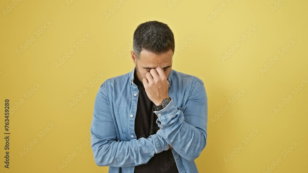 Stressed young hispanic man in denim shirt, isolated on yellow, rubbing tired eyes under strain from headache and fatigue Stressed young hispanic man in denim shirt, isolated on yellow, rubbing tired eyes under strain from headache and fatigue