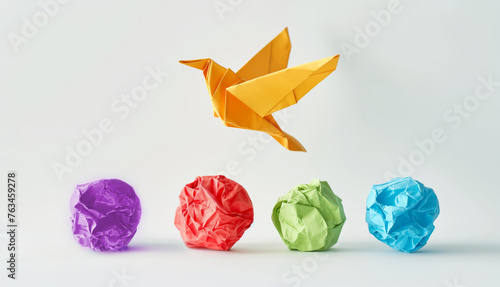 A photo of an origami bird flying over four differently colored crumpled paper balls on a white background, symbolizing creativity and imagination in business idea. photo