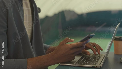 Slowmo of young Black male tennis coach sitting at table in front of laptop on second floor of spacious indoor tennis court and making phone call photo