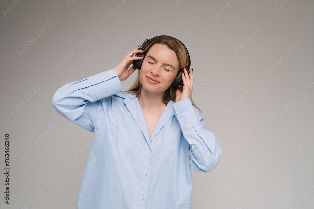 Enjoying the music with eyes closed. blonde woman in blue shirt touching her black headphones on her neck. Gray background studio shot Enjoying the music with eyes closed. blonde woman in blue shirt touching her black headphones on her neck. Gray background studio shot