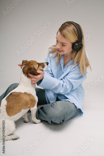 Blond woman hugging small dog Jack Russell terrier. Sitting on the floor listening to music. Blue jeans and shirt and gray background studio shot