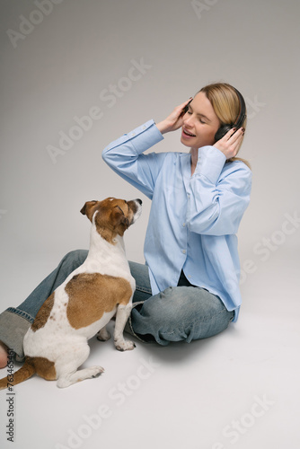 Girl and pet music lover. Blonde woman sitting on the floor with black headphones listening to song cute white dog looking at her with love. Studio shot. Gray and blue colors