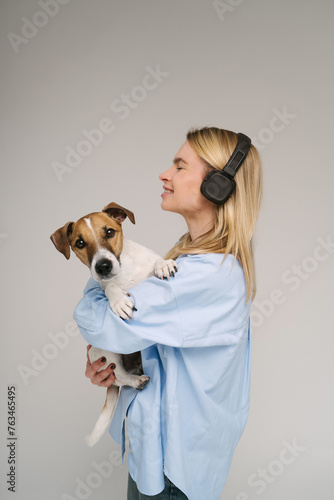 Enjoying music with closed eyes. Blonde young woman dog owner holding the pet. Funny Jack Russell terrier looking at camera. Studio shot grey and blue colors background