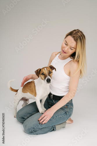 Blond woman sitting on floor and looking at small white dog Jack Russell terrier. Studio shot. Cute friends portrait. Dog owner care