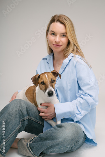 Smiling blonde woman in a blue shirt sits on the floor, the dog sitting on her laps. Funny cute friends studio portrait. Grey background. Vertical composition